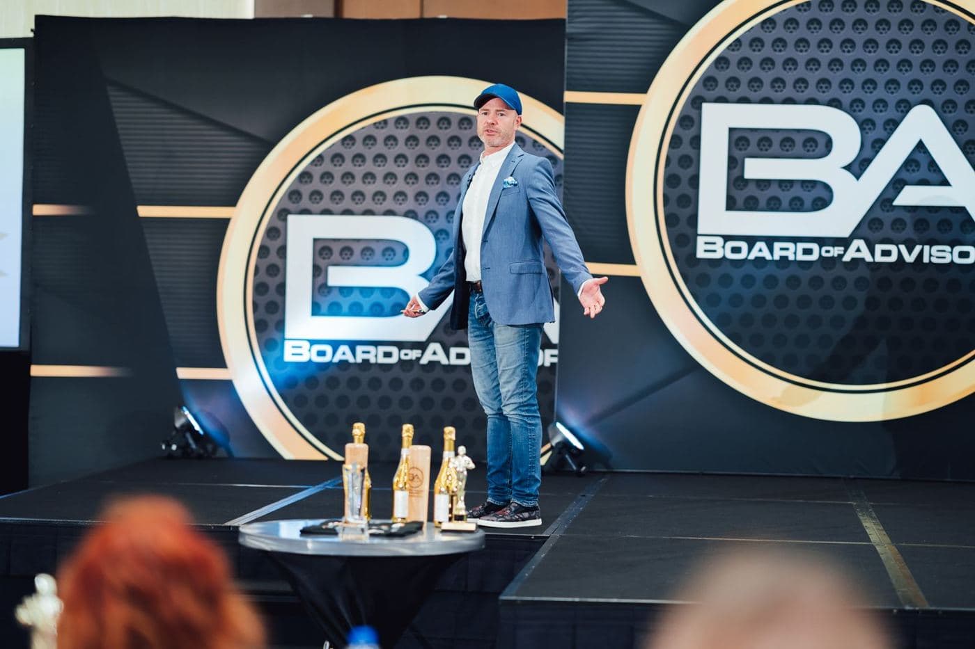 Mike Weiss on stage at a Board of Advisors event in a blue cap, arms open