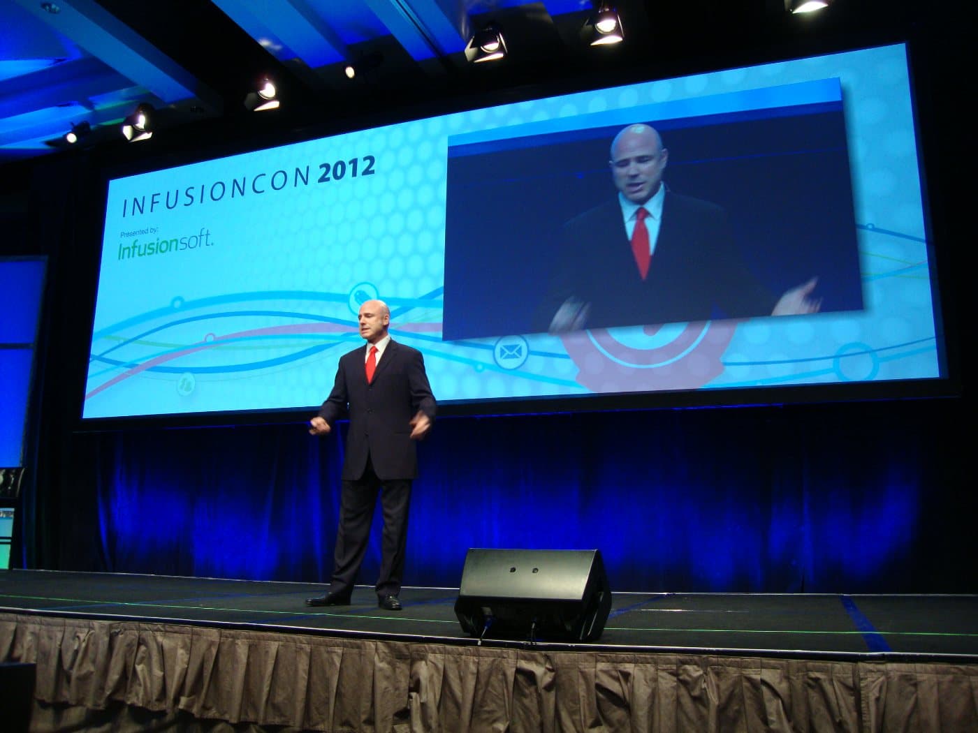 Mike Weiss speaking on the InfusionCon 2012 stage in a dark suit and red tie, shown on the big screen behind him