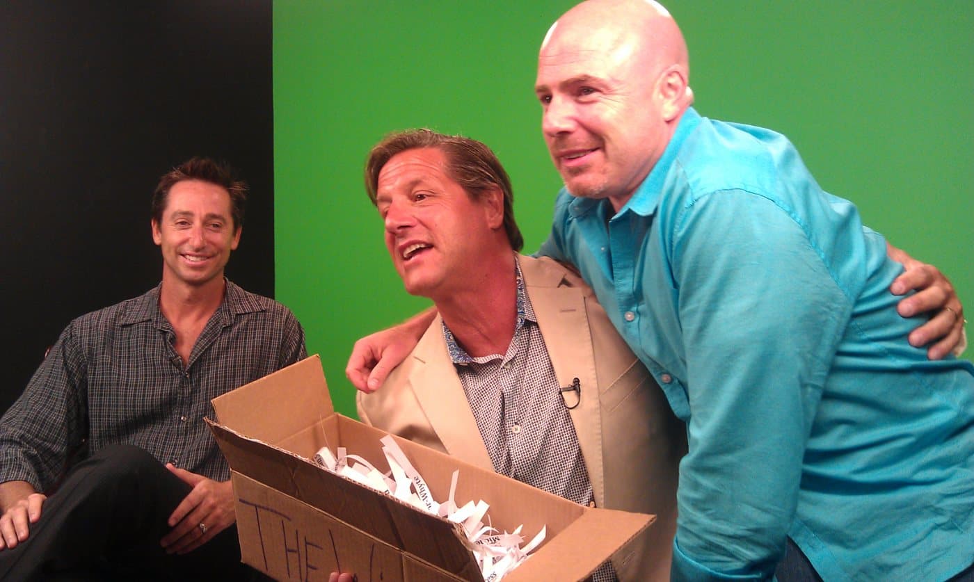 Mike Weiss hugging John Assaraf, seated, in front of a green screen