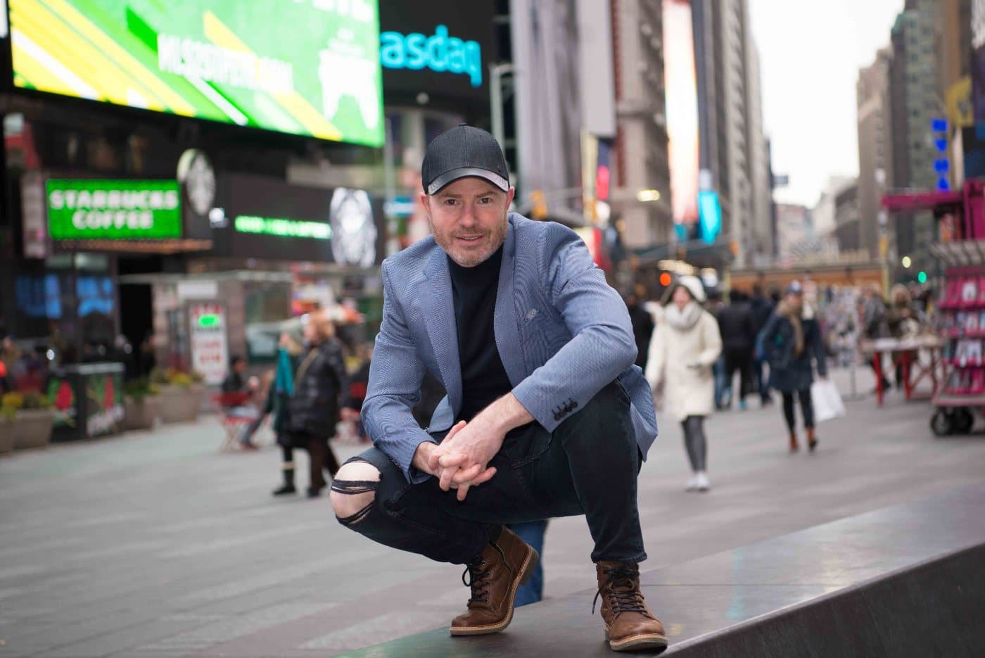Mike Weiss crouching on a ledge in Times Square, New York, in a dark cap and blue blazer